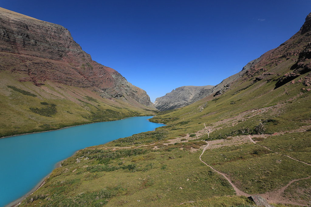 Cracker Lake Trail: One of the Most Beautiful Hikes in Glacier National Park 3 A view of Cracker Lake, bright turquoise, surrounded by green hillsides. You can see the Cracker Lake Trail on the side of the lake.