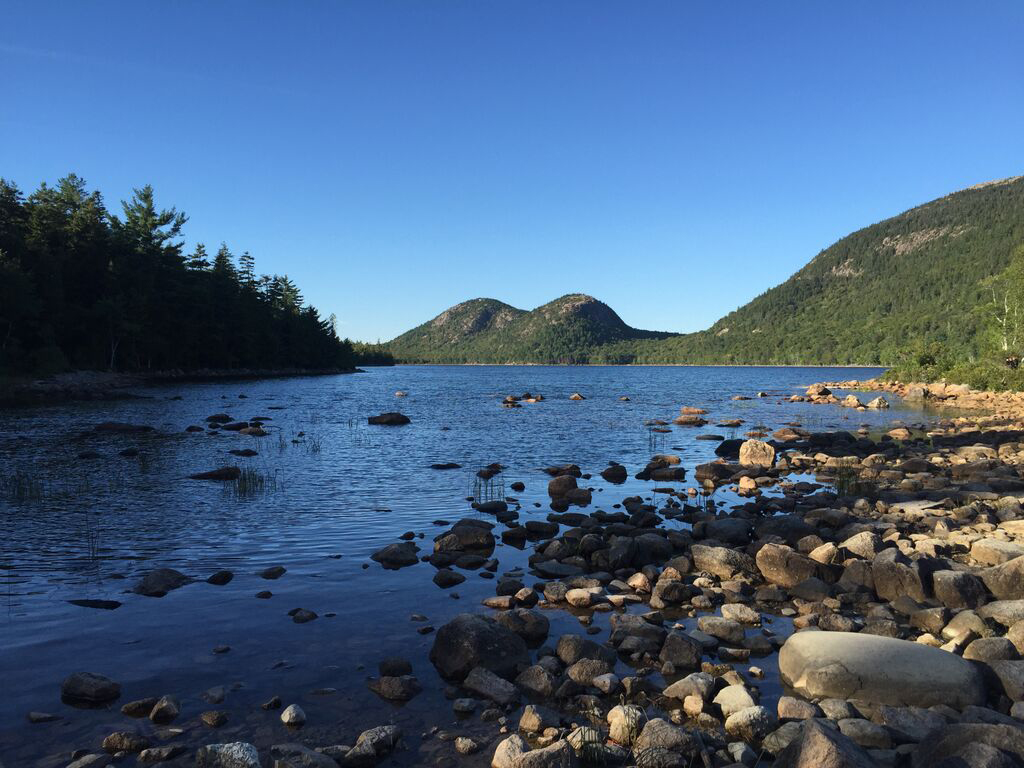 12 Things to Do in Acadia National Park That Will Take Your Breath Away 6 A blue pond with a rocky shoreline. On the other side are two green hills connected to each other with a small bowl in between. Visiting Jordan Pond is one of the best things to do in Acadia National Park.