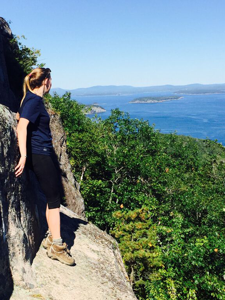 How to Hike the Heart-Stopping Precipice Trail in Acadia National Park 7 A female hiker clings to the side of a rockface above trees and ocean water. The Precipice Trail is one of the best things to do in Acadia National Park.
