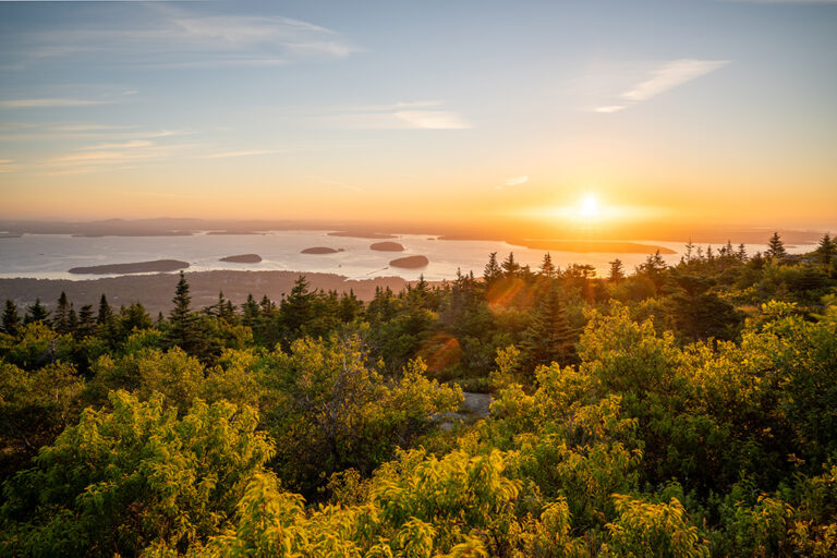 The sun rises above the ocean and a few islands. Acadia tops the list of 63 national parks in alphabetical order.