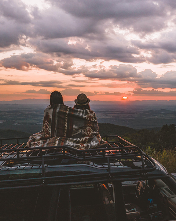 A couple watches the sunset while wrapped in a blanket and sitting on the roof of a van during a Shenandoah camping trip