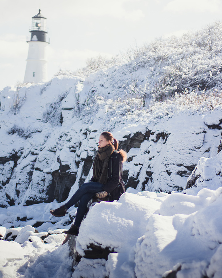 The Best Time to Visit Acadia National Park: When to See Fall Colors 16 A woman sits on rocks beneath a lighthouse. The entire landscape is covered in snow.