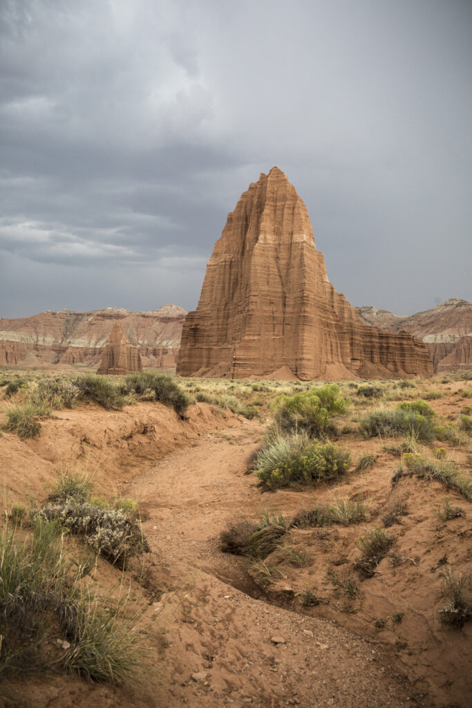 20 Best Hikes in Capitol Reef National Park 18 An orange trail leads to a tall rock monolith below gray skies
