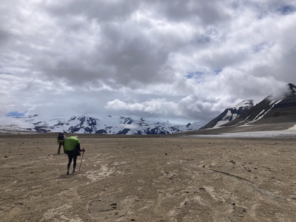 How to Get to Katmai National Park: Boats, Planes, and Bears, Oh My! 19 Two hikers with large backpacks walk toward snowy peaks across a barren landscape