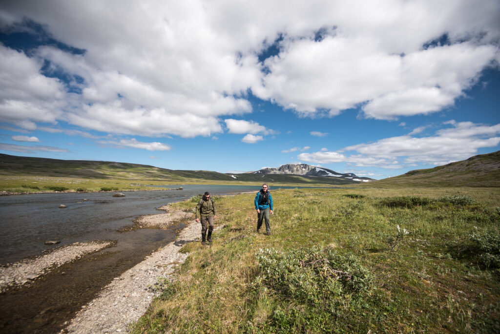 Where to Stay in Katmai National Park: Lodging, Campgrounds, and Backpacking 1 Two hikers walk toward the camera over a grassy area next to a river