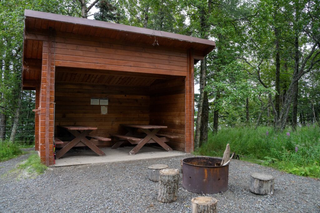 Where to Stay in Katmai National Park: Lodging, Campgrounds, and Backpacking 9 An open brown shelter with two picnic tables inside and a fire ring outside
